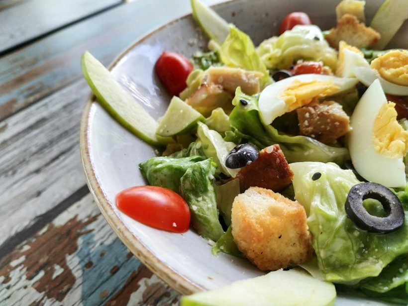 A close-up of a Caesar salad featuring fresh lettuce, eggs, croutons, and olives on a rustic table.