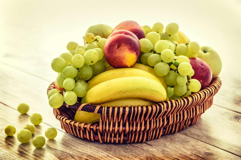 A colorful wicker basket filled with fresh grapes, bananas, and apples on a wooden table.